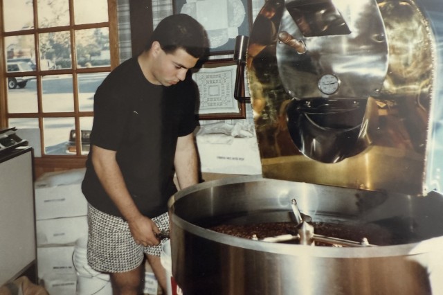 Teenage boy in black t-shirt and black & white shirts turning beans in a large coffee roaster neat a window inside a bakery.